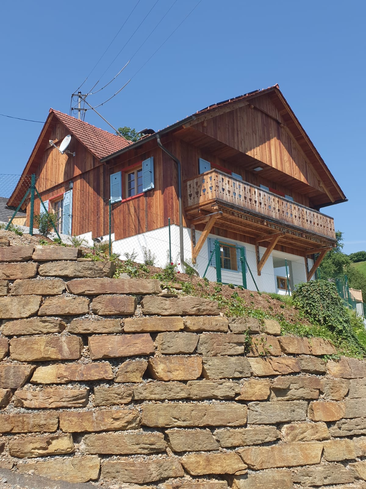 The well-constructed villa features a charming wooden exterior and a sloped roof. A balcony with wooden railing is visible on the upper level, providing outdoor space. The building is set against a clear blue sky, with surrounding greenery and a stone wall at its base.