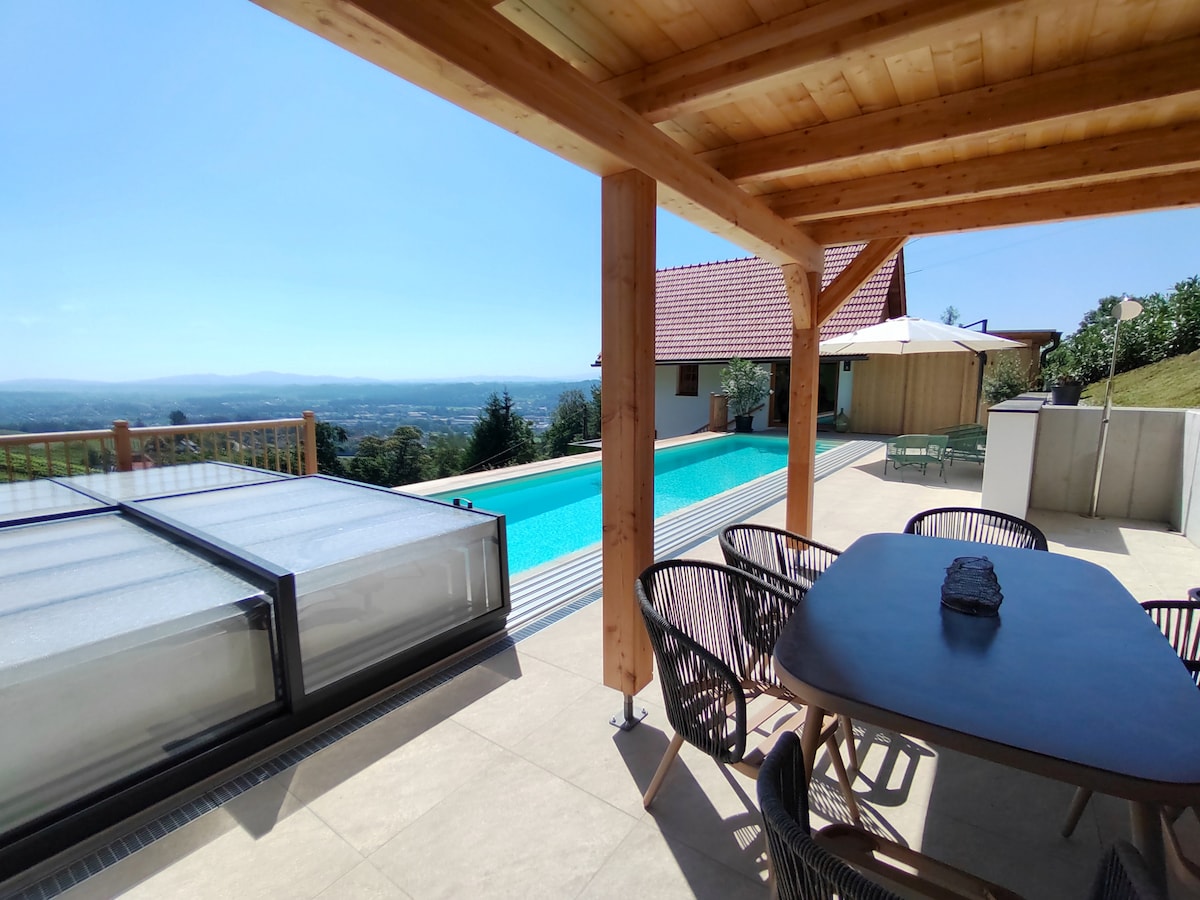 A spacious outdoor terrace offers a dining set with multiple chairs, overlooking a large pool. A sun umbrella provides shade, and a wooden structure supports the roof. The surrounding landscape features panoramic views of vineyards and hills under a clear blue sky.