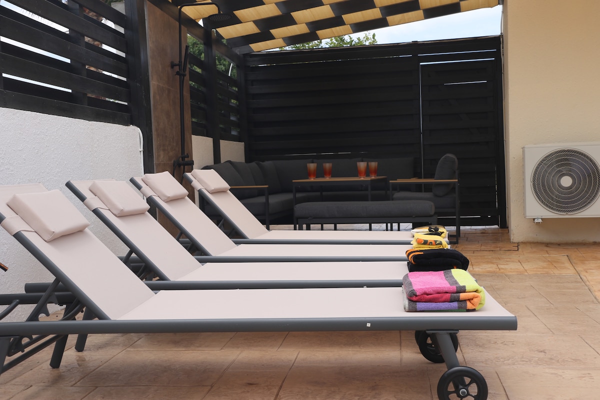A sun terrace is featured with four lounge chairs arranged in a row. Bright towels in various colors are neatly placed on the ground. A shaded seating area is visible in the background, accompanied by two glasses on a table, providing a relaxed outdoor space.