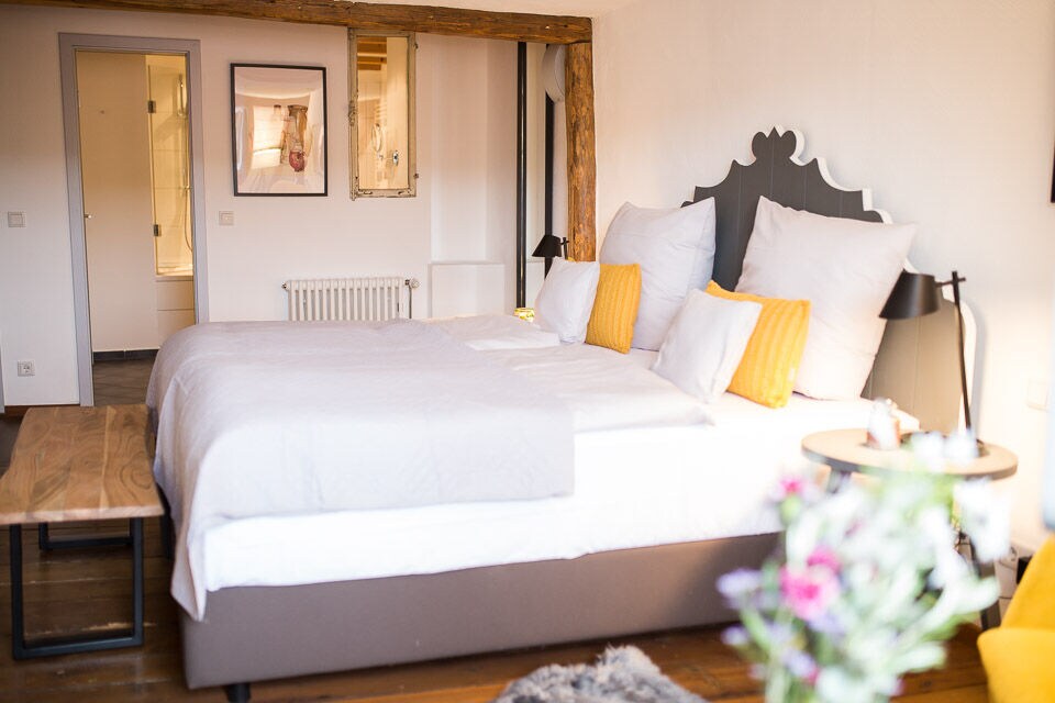 A well-appointed room features a spacious bed dressed in crisp white linens, complemented by yellow accent pillows. Wooden beams add character to the space, while a small table and soft rug create a cozy environment. A modern bathroom is visible in the background.