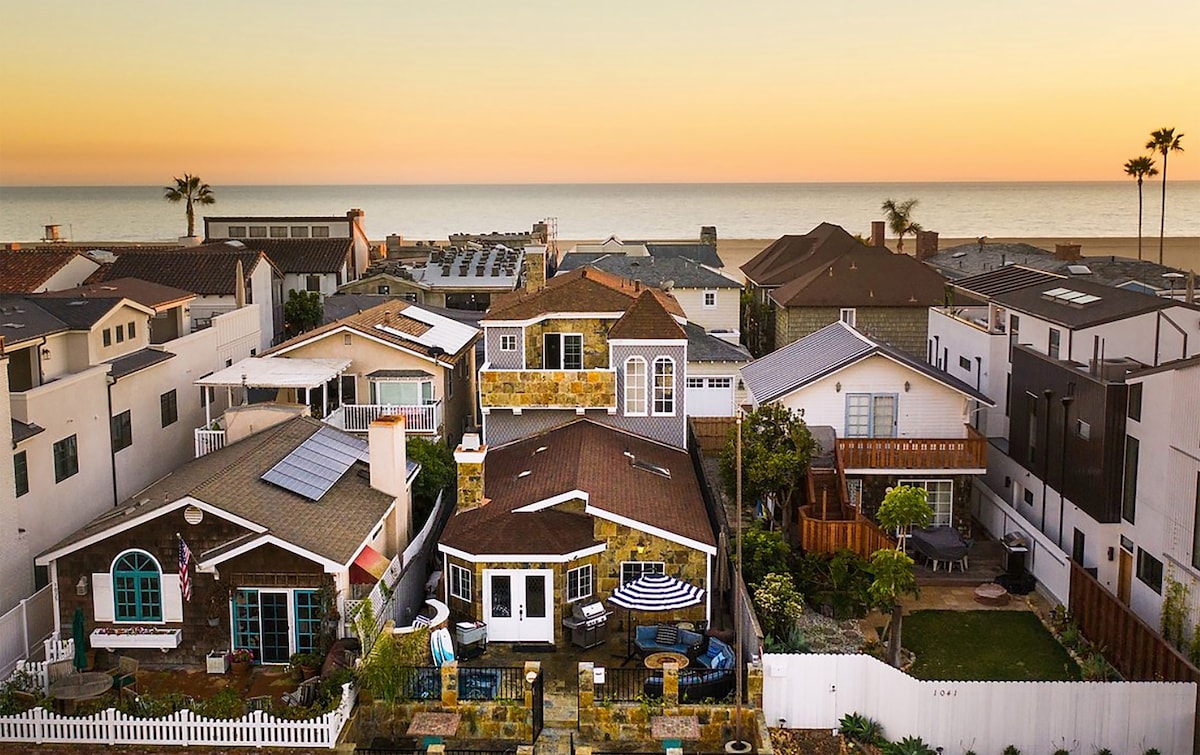 An aerial view captures a charming coastal neighborhood at sunset, where homes feature a mix of architectural styles. The calm ocean glistens in the background, and palm trees sway gently, adding to the serene atmosphere.