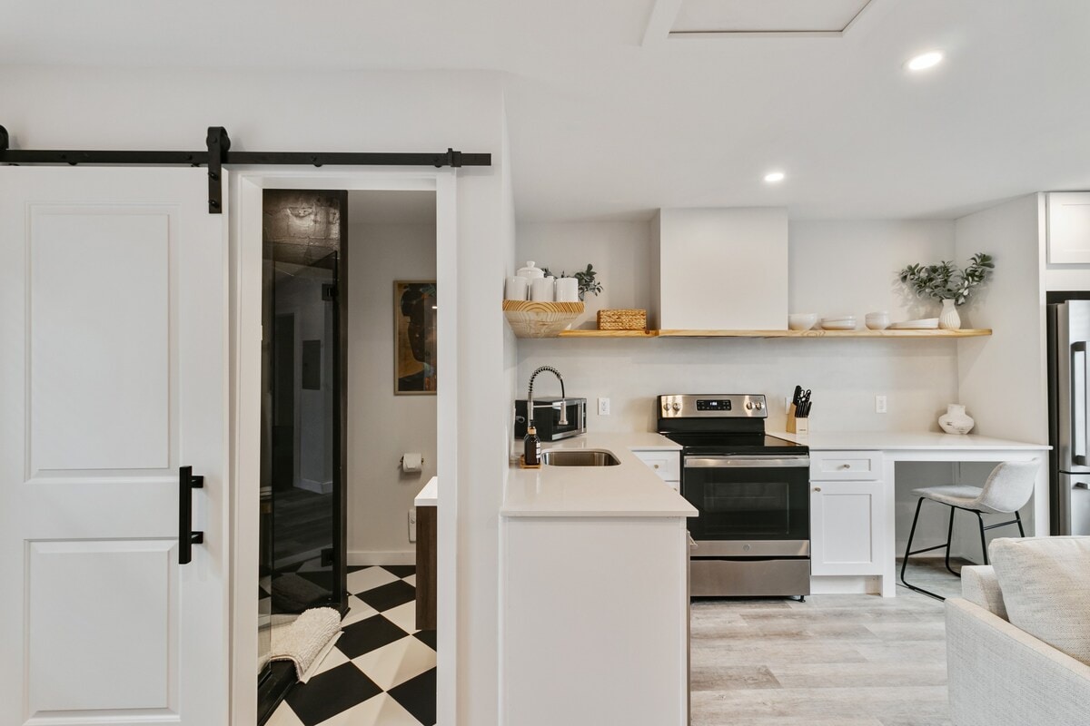 The modern kitchen is visible, featuring sleek appliances and minimalist shelving stocked with dishware. A barn-style sliding door leads to the bathroom area. The open layout allows for easy movement between spaces, highlighted by contemporary flooring and stylish decor.