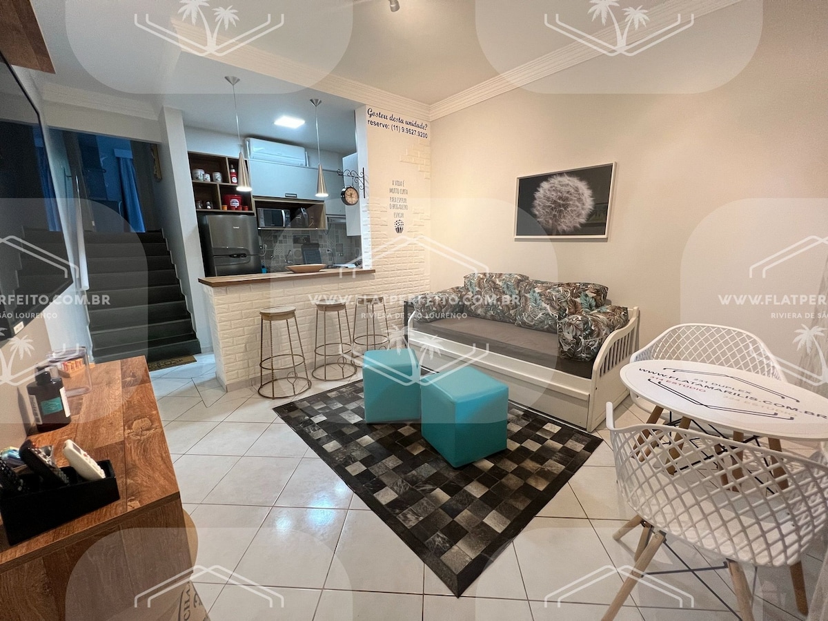 A comfortable living area features a modern sofa adorned with patterned cushions. A coffee table in a subtle turquoise hue is positioned in front. Barstools are situated near a kitchen counter, and a dining table is set for four, creating a welcoming space for relaxation.