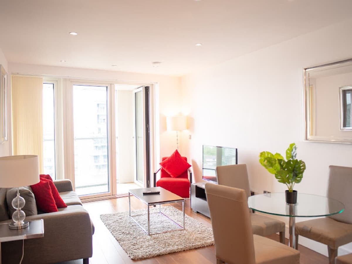 A modern living/dining area features a gray sofa and a bright red armchair. A glass coffee table is centered on the wooden floor, with a round dining table for four visible. Large windows open to a small balcony, allowing for ample natural light.