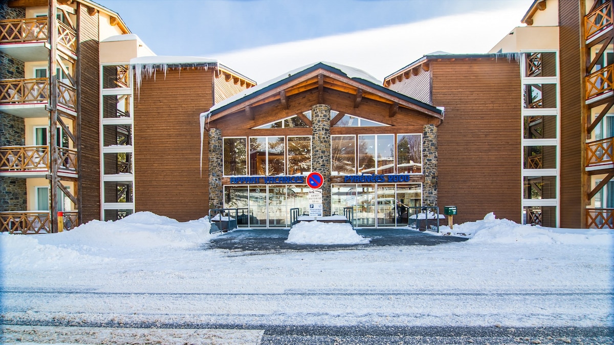 The exterior of a rustic building features wooden and stone elements, complemented by a peaked roof. Snow covers the ground, and the entrance displays large glass doors. Balconies with wooden railings are visible on upper floors, offering a cozy mountain retreat feel.