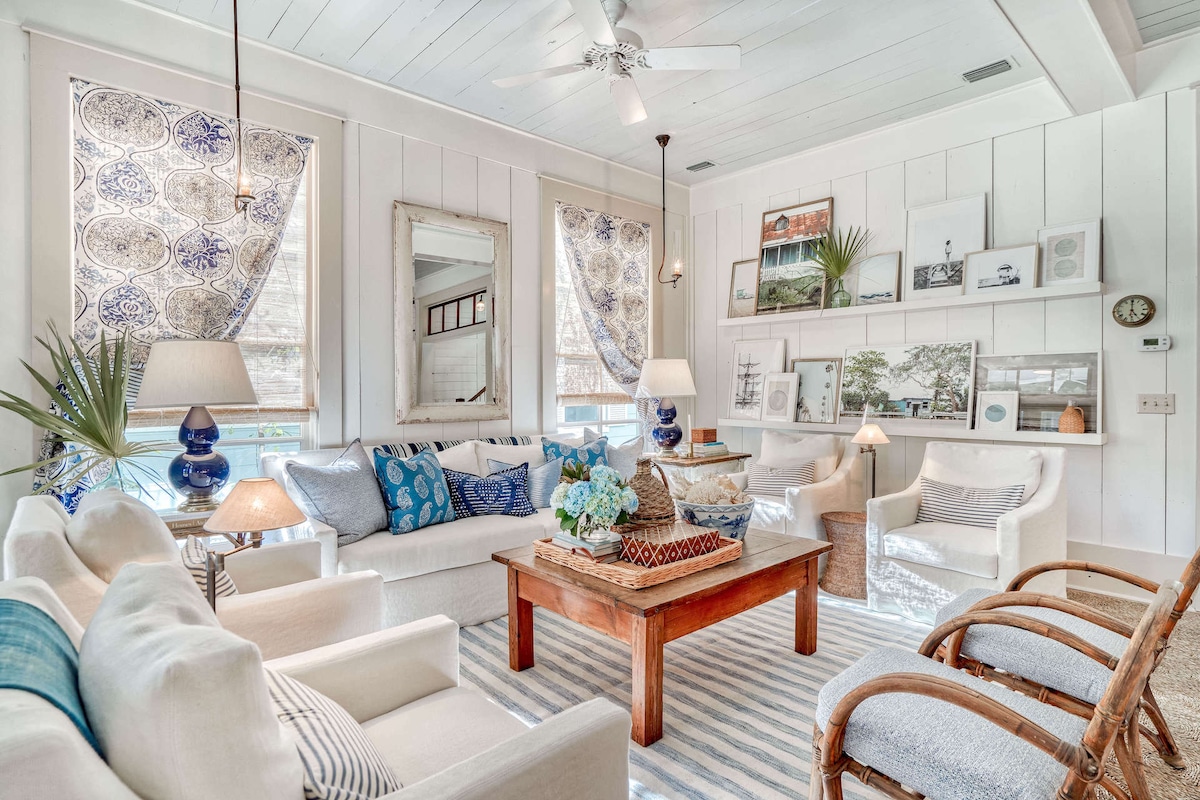 A light-filled living area features white sofas adorned with blue and patterned throw pillows. A wooden coffee table is central, surrounded by woven chairs. Natural light streams through patterned window coverings, highlighting the coastal decor including framed photographs and houseplants.