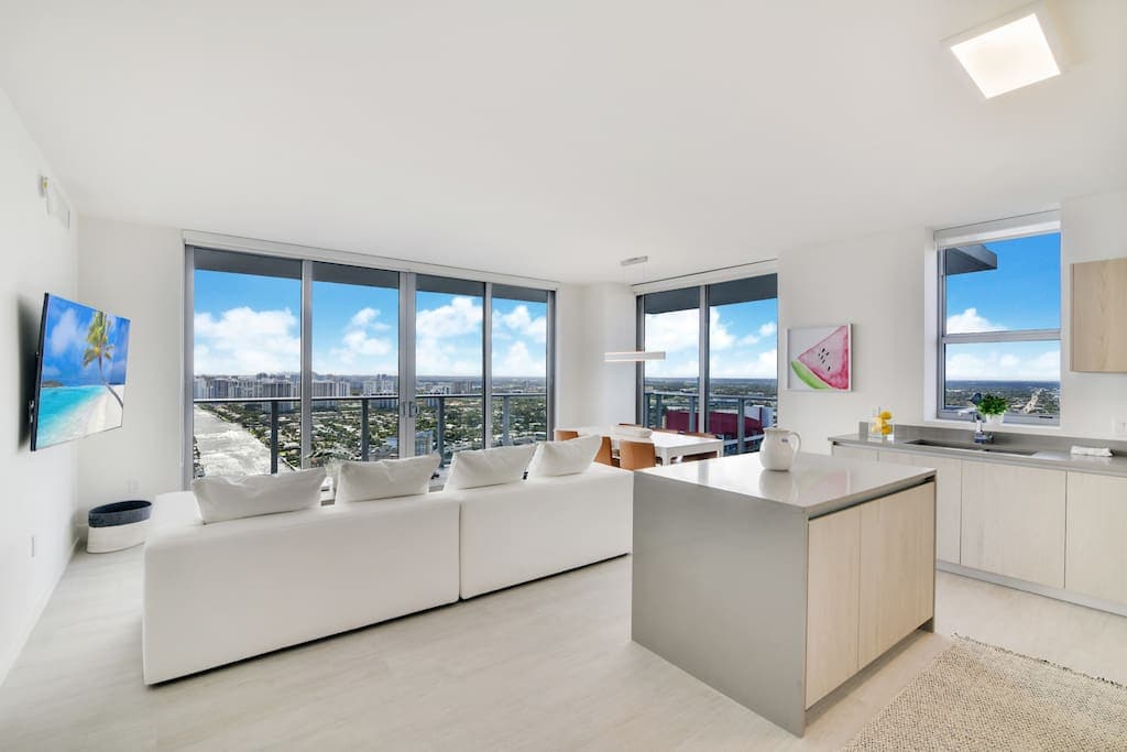 A modern living area features large floor-to-ceiling windows, offering expansive views of the surrounding area. A sleek kitchen island with a stainless steel finish complements the contemporary design. A sectional sofa in the seating area provides a comfortable space for relaxation.