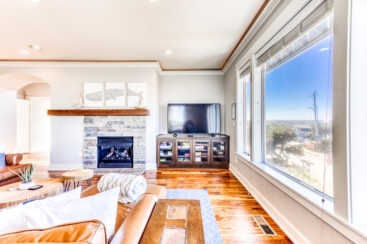 A bright living area features large windows offering views of the outdoors. A contemporary television is mounted above a stylish stone fireplace, and a wooden entertainment unit is stocked with games and media. Warm wooden flooring complements the inviting atmosphere.