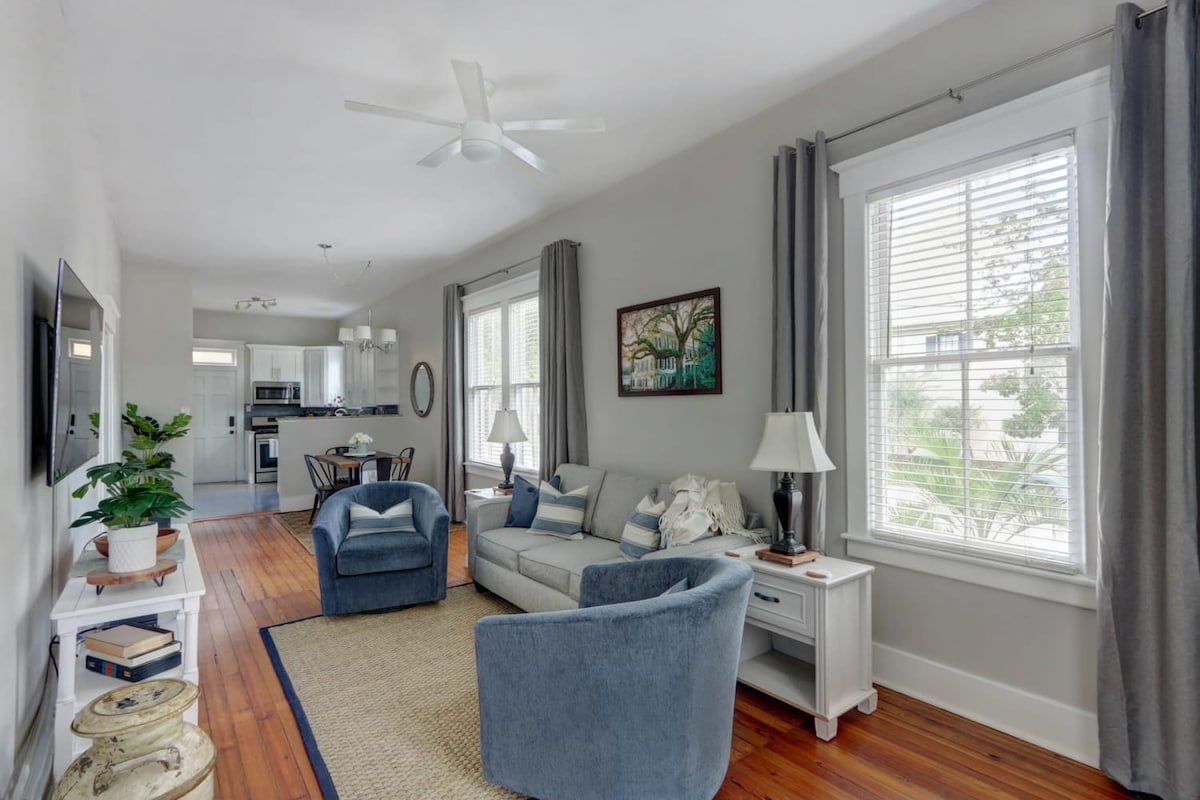 A welcoming living area features a gray sofa and two blue chairs, complemented by a light rug. Natural light streams through large windows adorned with sheer curtains. A dining table for four is visible in the adjacent space, enhancing the open layout.