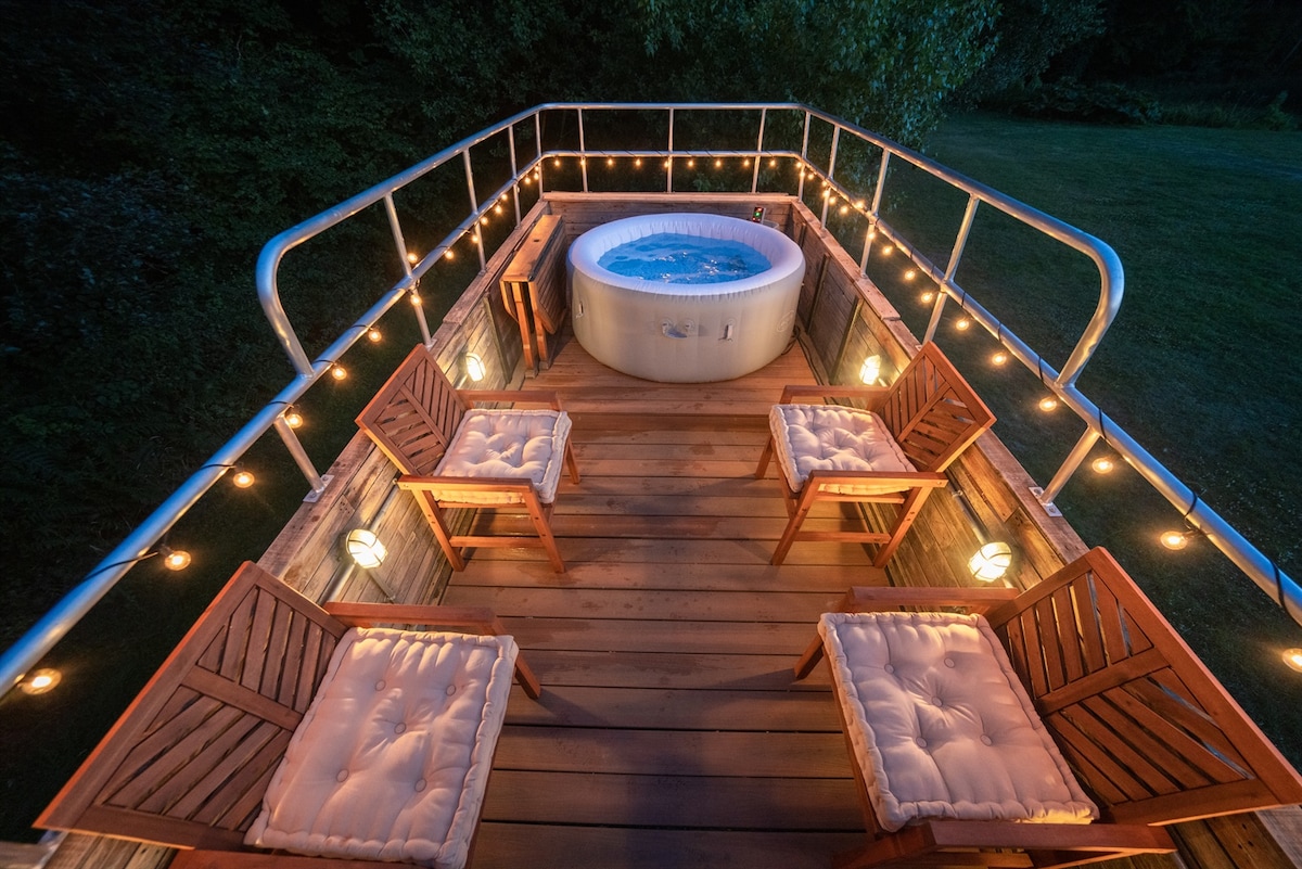 The rooftop area features a circular hot tub surrounded by wooden decking. Five wooden chairs are positioned around the hot tub, offering seating. Soft lighting is provided by string lights along the railing, creating a warm glow in the evening.