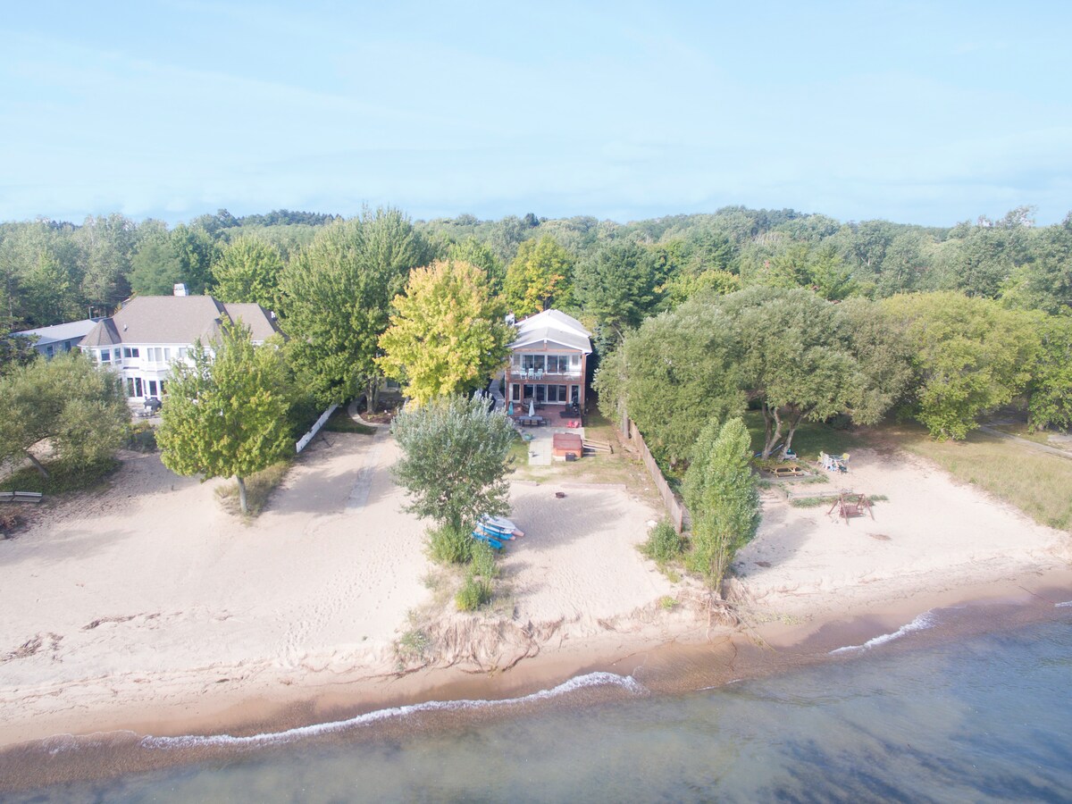 The lakefront home sits on a sandy beach surrounded by lush greenery. A spacious outdoor area features seating and pathways leading to the water, creating an inviting space for relaxation. The serene setting is complemented by the gentle waves lapping at the shore.