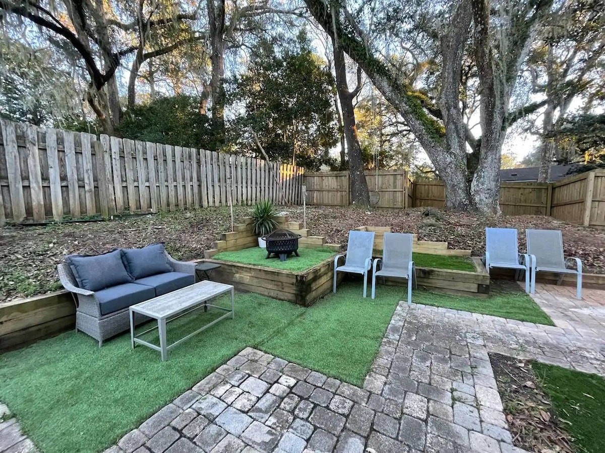 A spacious backyard is enclosed by a wooden fence and shaded by mature trees. Comfortable seating is arranged around a fire pit on a mix of grassy areas and stone pavers, providing an inviting outdoor space for gatherings.