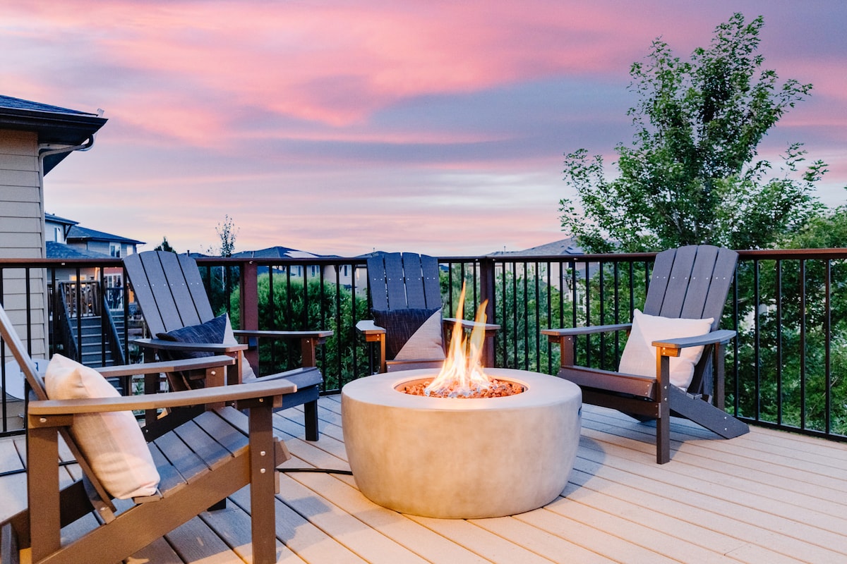 A spacious outdoor deck features a circular gas fire pit surrounded by comfortable wooden chairs. The area is enhanced by a colorful sunset sky, providing a serene atmosphere for relaxation and gatherings.
