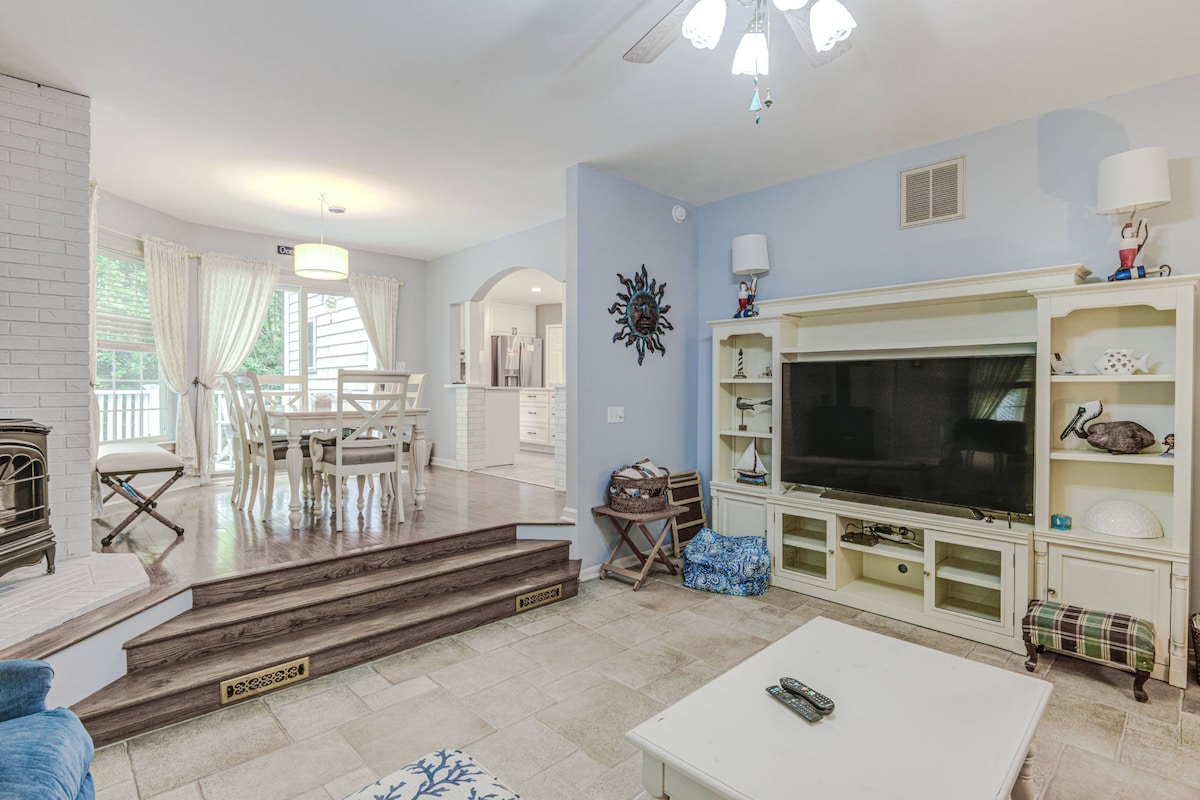 A spacious living area is highlighted by a light blue wall and a white entertainment center. Steps lead to a cozy seating area, featuring a comfortable sofa and decorative accents. Natural light fills the room, enhancing the view of the adjoining dining space and kitchen.