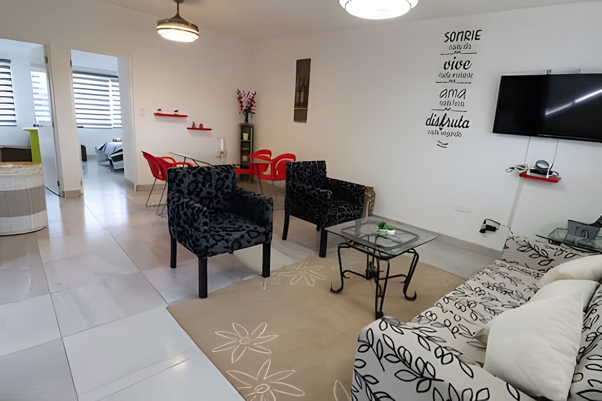 A modern living space is displayed with patterned black chairs arranged around a glass table. A light-colored couch adds contrast, and decorative wall art features uplifting phrases. Natural light brightens the room, and soft area rugs provide comfort underfoot.