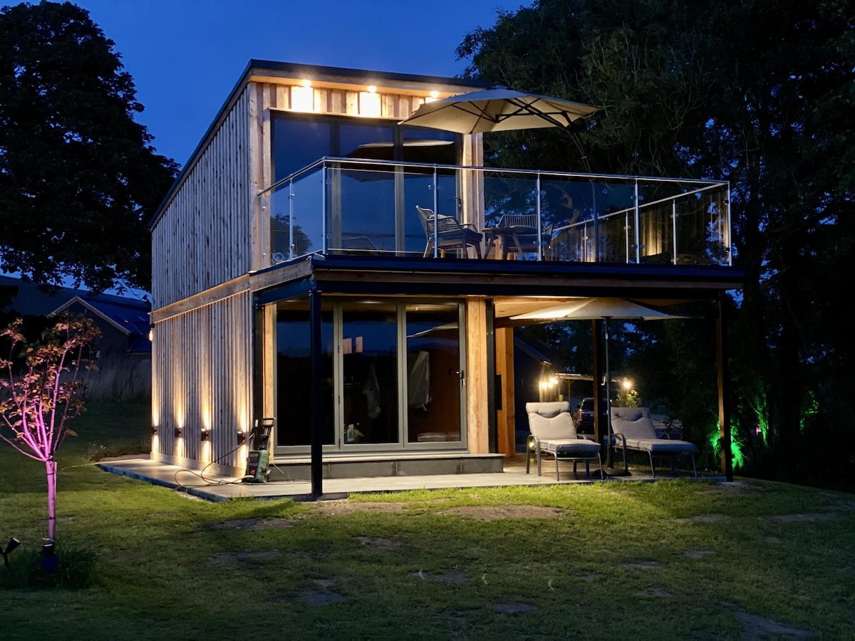 The lodge's modern exterior is spotlighted at night, showcasing wood paneling and large glass windows. A balcony with outdoor seating is visible, accompanied by a sleek umbrella. Ambient lighting highlights the landscape, creating a serene evening atmosphere.