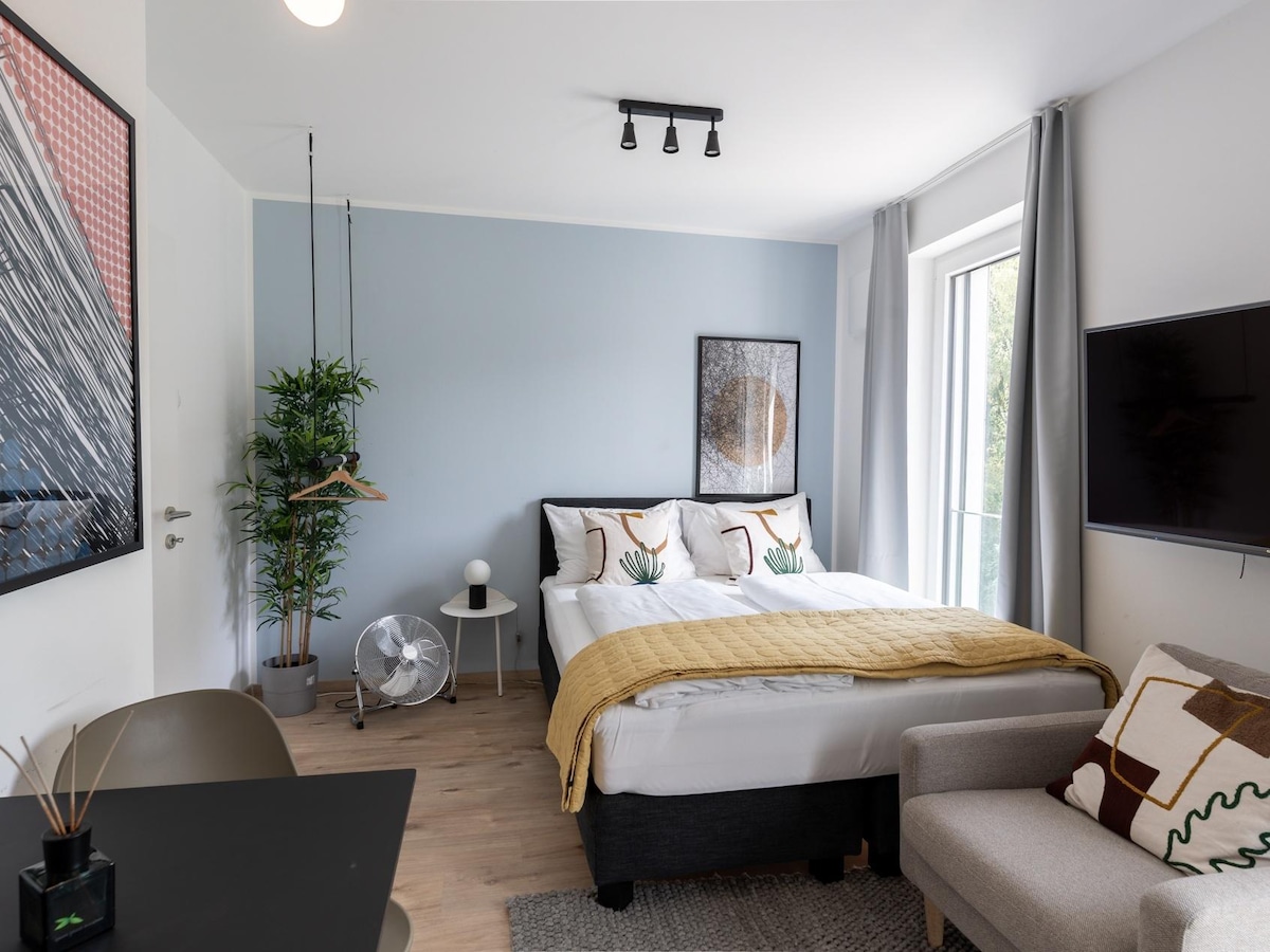 A modern room features a spacious bed adorned with patterned pillows and a warm throw blanket. A small sofa sits nearby, complemented by a wall-mounted TV. Natural light enters through a window with light curtains, while decorative artwork adds character to the space.