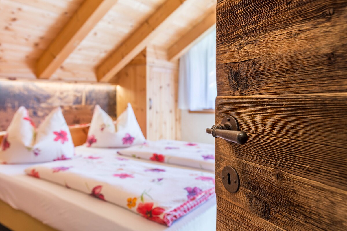 A glimpse into a cozy bedroom is presented, featuring a spacious bed adorned with floral-patterned linens and decorative pillows. Wooden beams and paneling create a warm ambiance. Natural light filters through a window, adding brightness to the inviting space.
