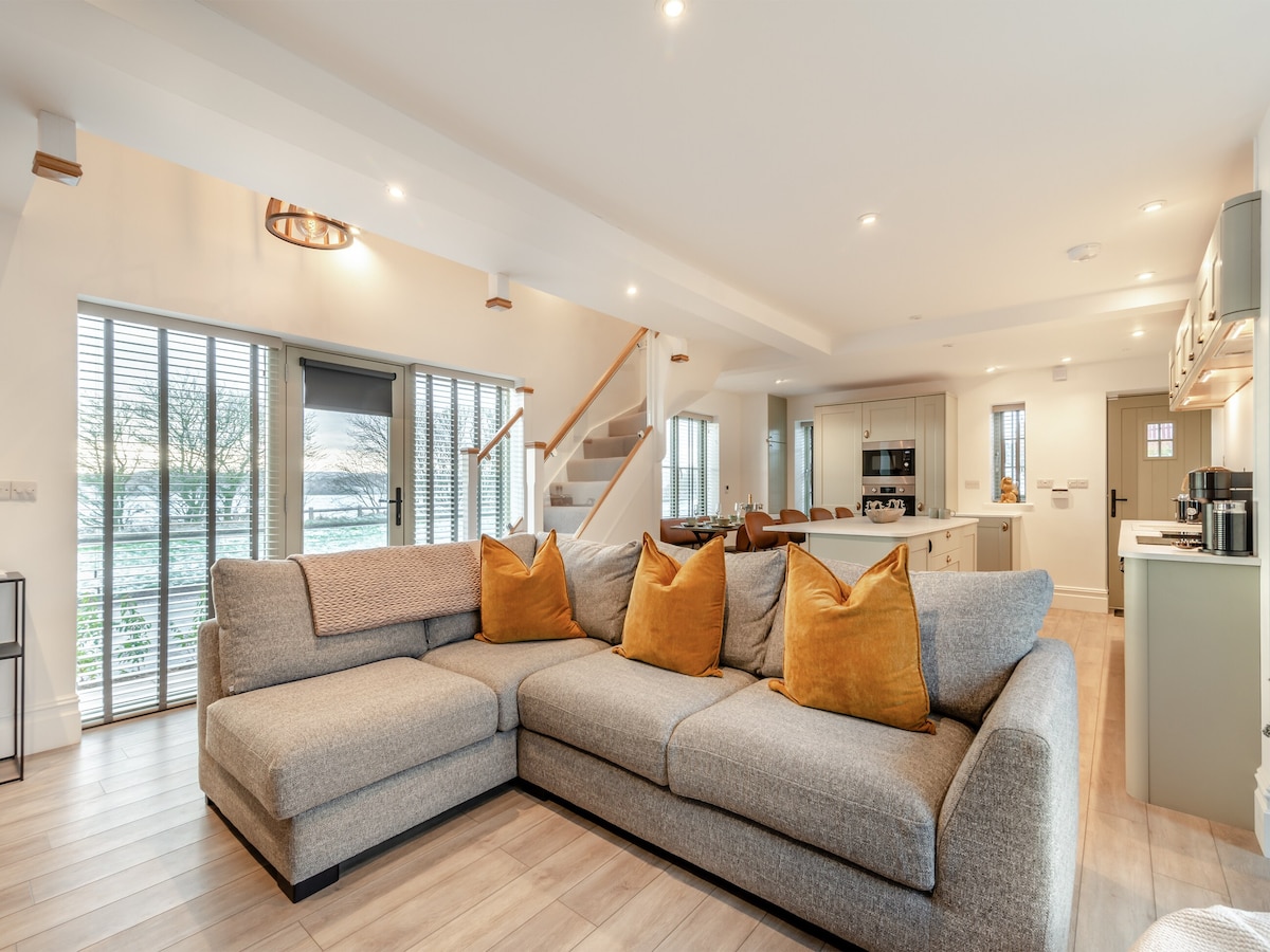 An open-plan living area features a large, L-shaped gray sofa adorned with mustard-colored cushions. A kitchen can be seen in the background, accompanied by a dining area. Glass doors provide ample natural light and beautiful views of the surrounding landscape.