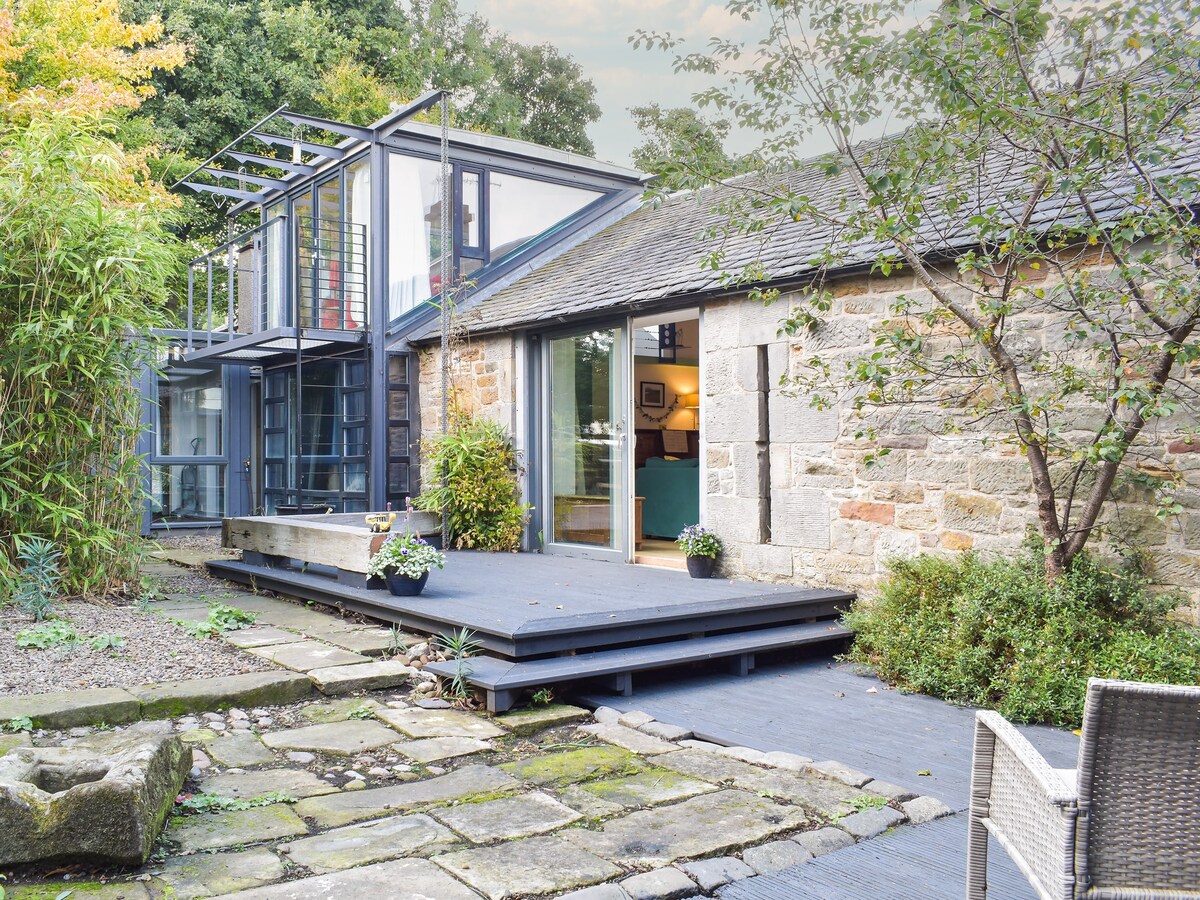 The exterior of the stone-built cottage features a modern extension with large glass panels. A wooden deck provides access to the garden, surrounded by lush greenery. A comfortable chair is positioned nearby, offering a spot to relax and enjoy the surroundings.