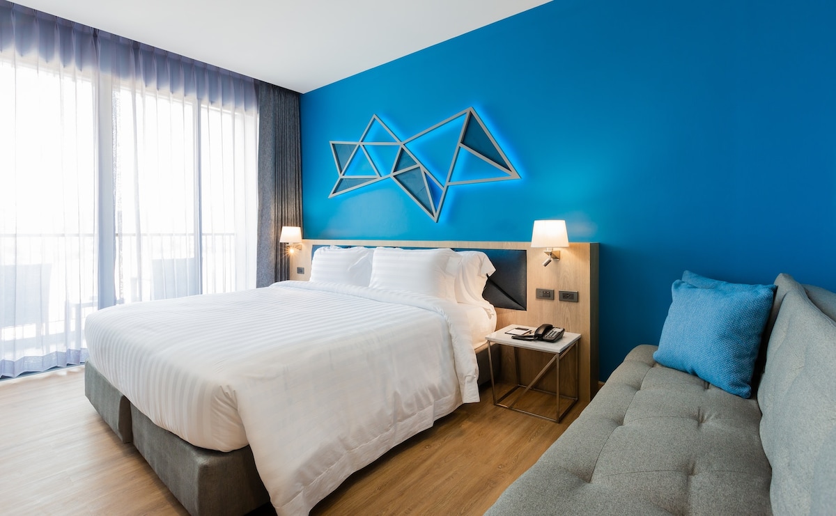A modern guest room features a spacious bed with crisp white linens against a vibrant blue wall. A light gray sofa complements the decor. Natural light floods the space through floor-to-ceiling windows, adorned with sheer drapes, creating a bright and open atmosphere.
