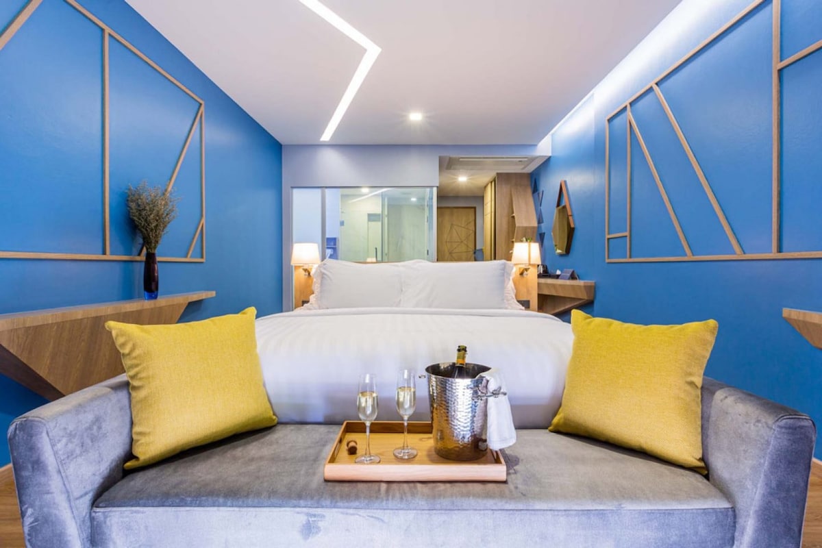 A modern room features a king-size bed with crisp white linens. Two yellow decorative pillows contrast with the gray sofa in the foreground. A wooden tray holds two glasses and a champagne bucket. Blue accent walls and soft lighting create a relaxed ambiance.