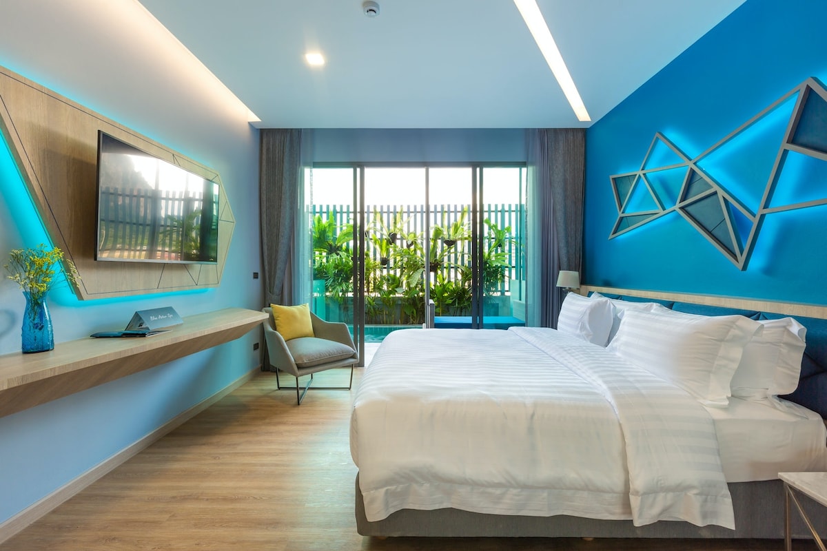 A spacious bedroom is highlighted by a double bed covered with crisp white linens. A large LED TV is mounted on the wall above a wooden console. Natural light filters through the glass doors opening to a private balcony, adorned with greenery.