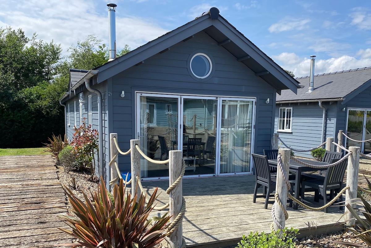 A detached beach house is showcased with a charming blue exterior. Large bifold doors provide an inviting entrance to a spacious wooden deck, furnished with a dining table and chairs. Native plants surround the area, enhancing the connection to the natural surroundings.