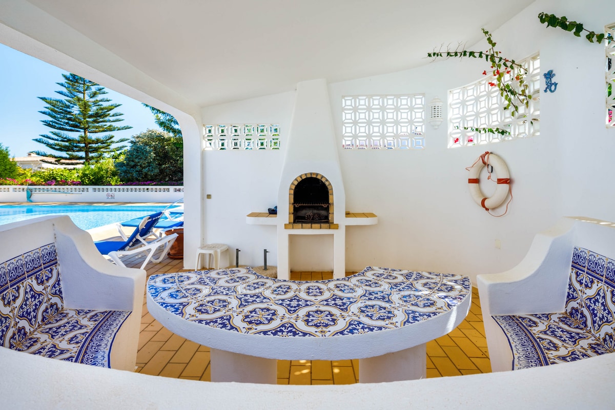 A covered BBQ area is shown, featuring a tiled table with decorative blue patterns and built-in seating. The area is bathed in natural light, with a lifebuoy mounted on the wall. The swimming pool is visible in the background, framed by sunbeds and greenery.