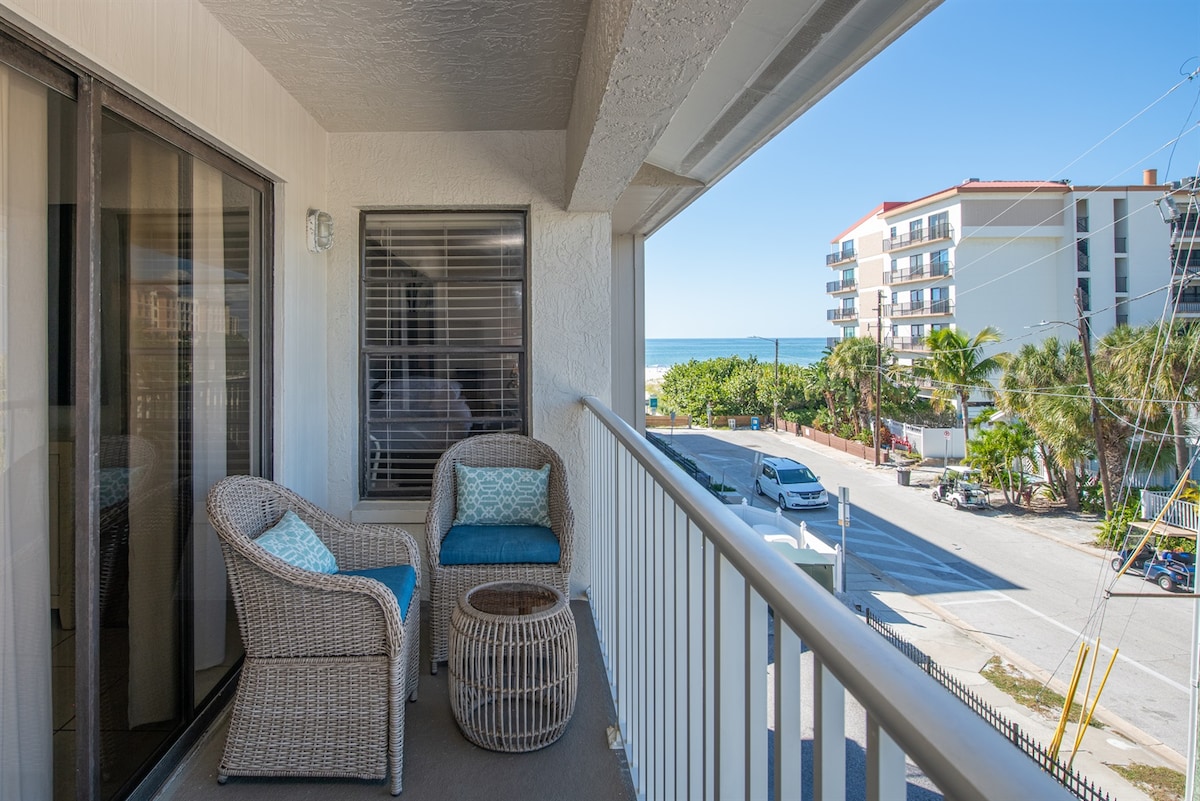 A private balcony features two wicker chairs with blue cushions and a round table. The view includes the street and a glimpse of the beach, surrounded by palm trees and nearby buildings. Natural light fills the space, enhancing the serene outdoor setting.