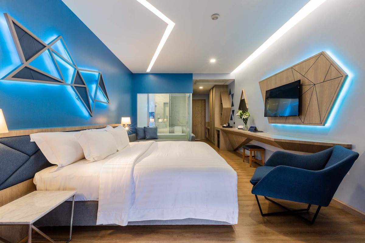 A modern guest room features a spacious bed with crisp white linens, complemented by blue accent walls and geometric decorative elements. A sleek work desk is positioned beside a blue accent chair. The bathroom is visible through a glass partition, providing an open feel.
