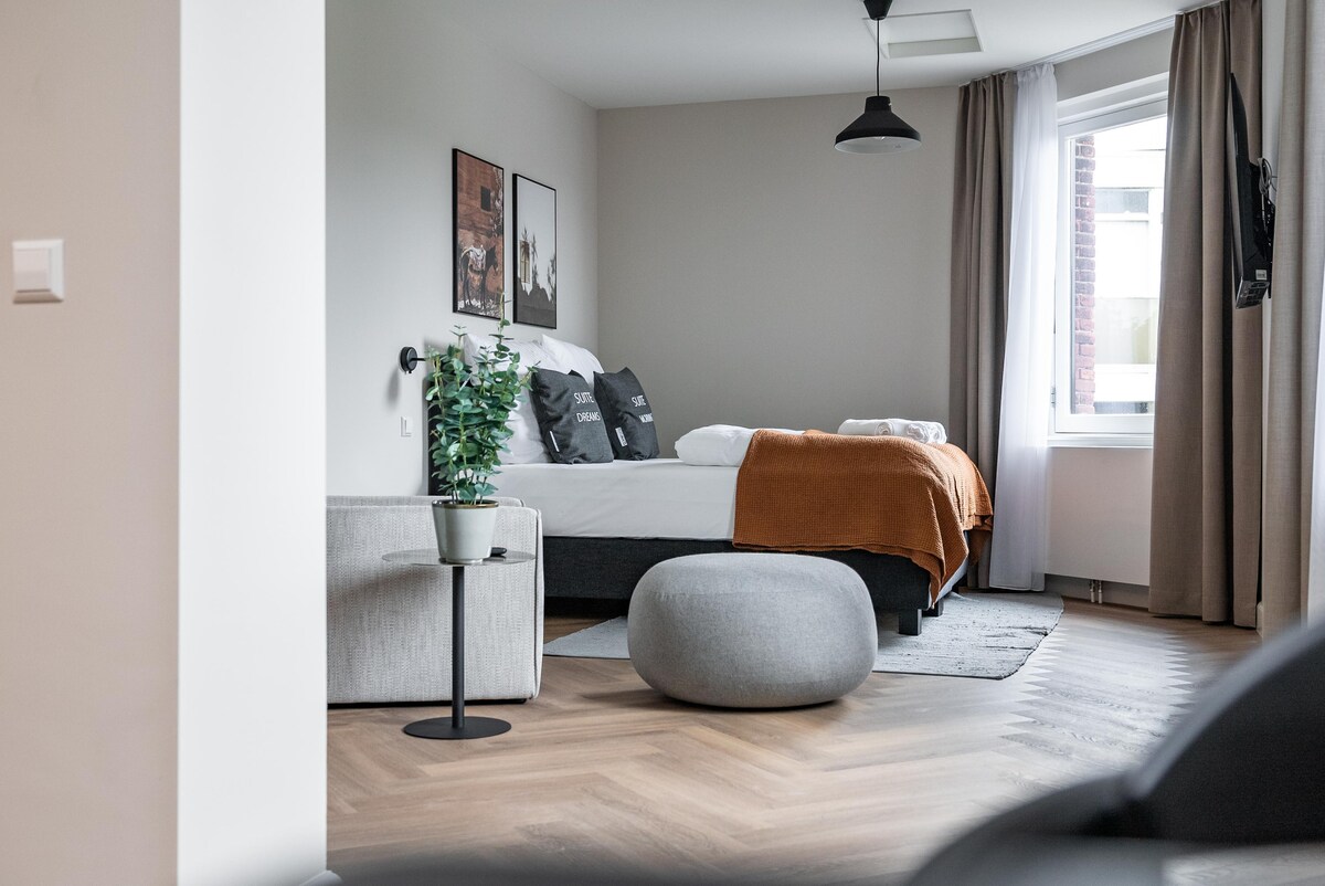 A spacious room features a comfortable box-spring bed dressed in neutral tones, accompanied by decorative pillows. A cozy orange throw is draped over the bed. The living area includes a soft gray seating option and a round ottoman. Natural light enters through a large window with sheer curtains.