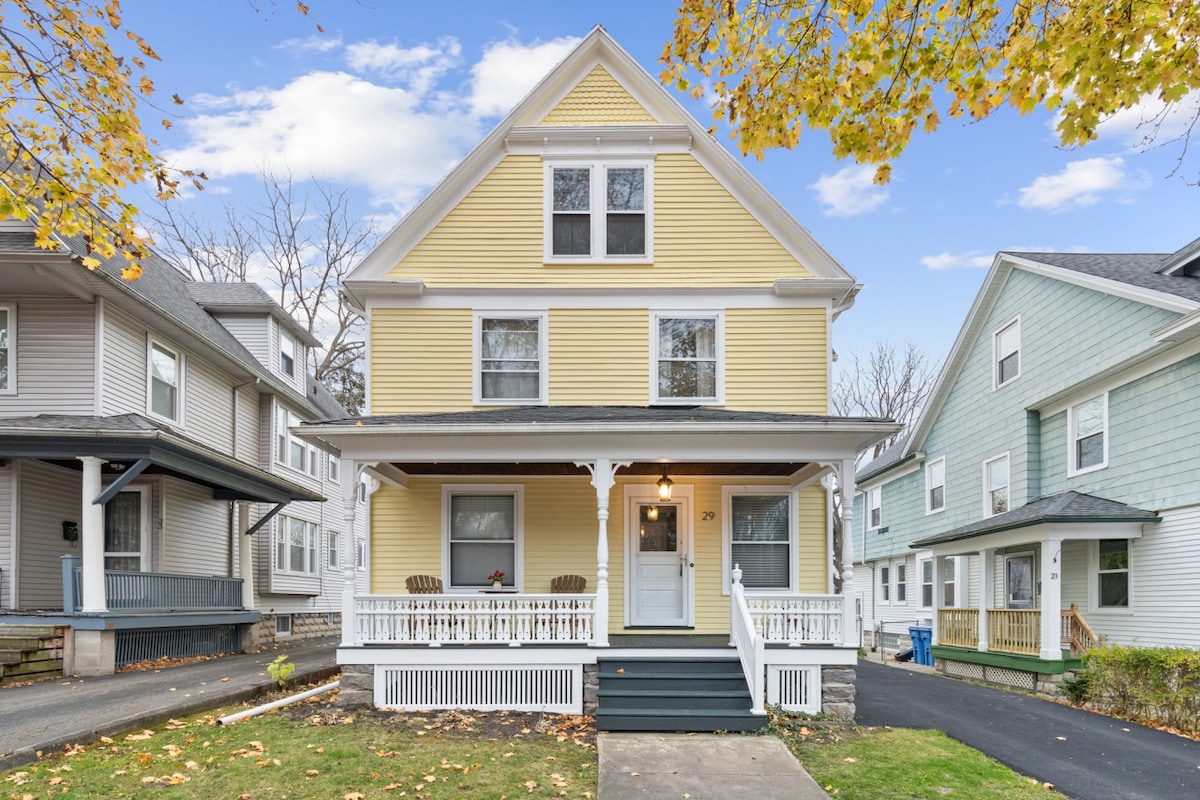 A charming three-story yellow house is presented, featuring a welcoming front porch with seating arrangements. Large windows on each level allow natural light to fill the interior. Surrounding homes are visible, indicating a residential neighborhood with a calm atmosphere, highlighted by autumn leaves.