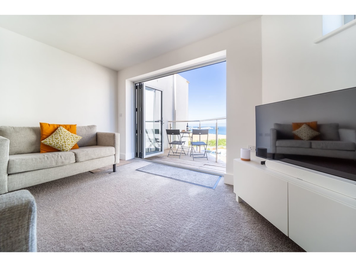 A spacious living area features a comfortable sofa adorned with an orange cushion, complemented by a sleek white entertainment unit with a television. Bi-fold doors open to a private terrace, showcasing views of the sea and outdoor seating for two.