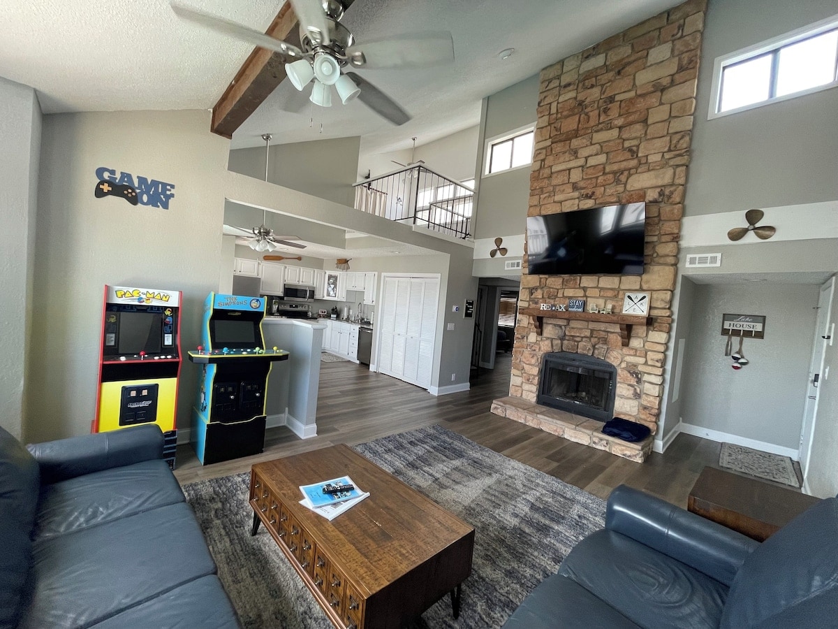 An open living area features a stone fireplace and large windows allowing natural light. A game corner includes vintage arcade machines. Comfortable seating is arranged around a wooden coffee table, with a view of the kitchen and loft area in the background.