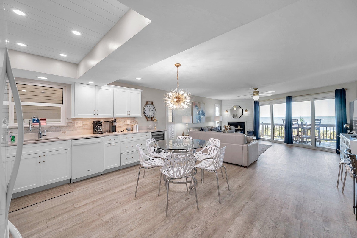 An open-concept space showcases a fully equipped kitchen with white cabinetry and a dining area featuring a round table surrounded by four white chairs. A cozy living area is visible, with large glass doors framing ocean views, allowing natural light to brighten the room.