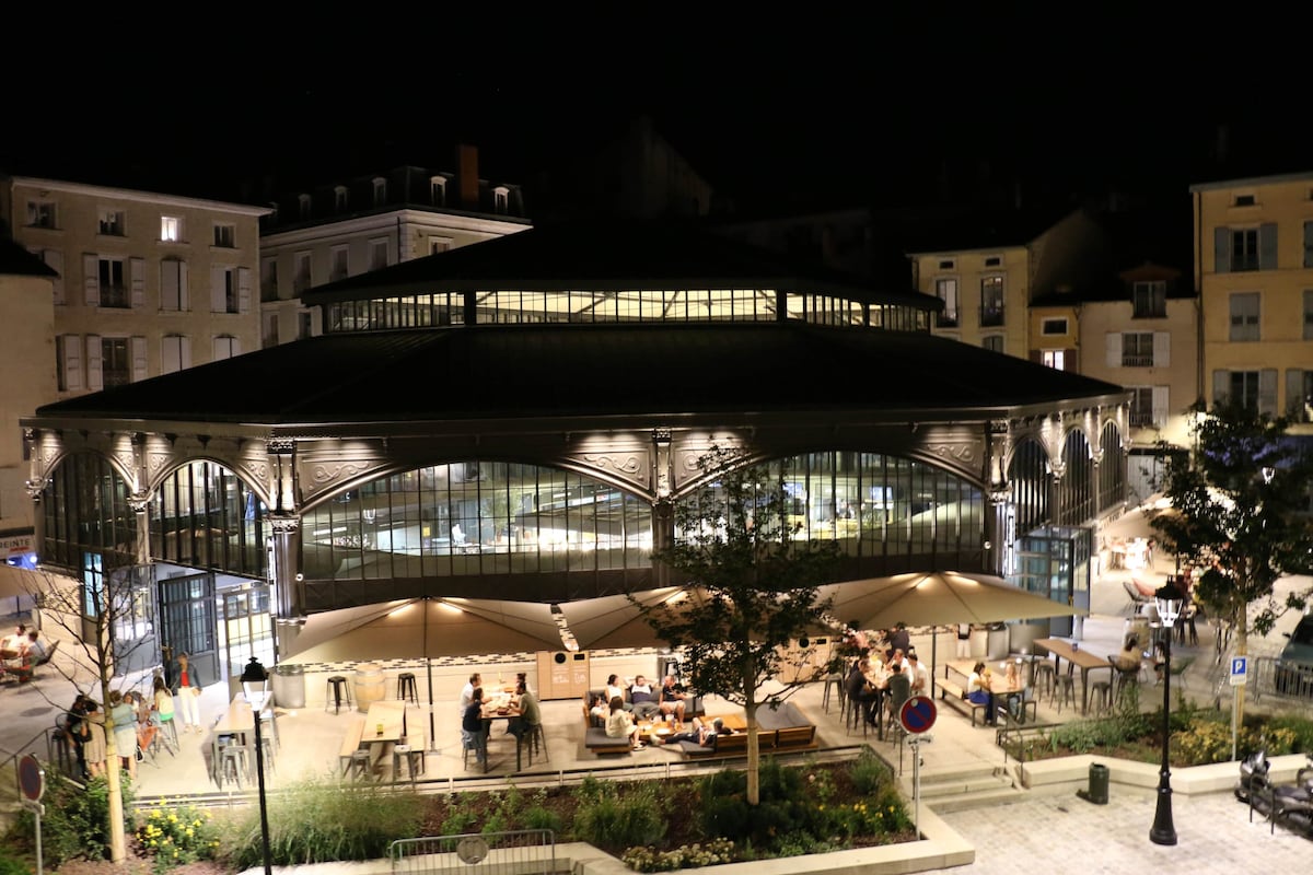 The Covered Market is illuminated at night, showcasing its unique architectural design with large glass sections. Outdoor seating areas are visible, with patrons enjoying the ambiance. Surrounding buildings contribute to the vibrant atmosphere, while modern light fixtures enhance the inviting appearance.