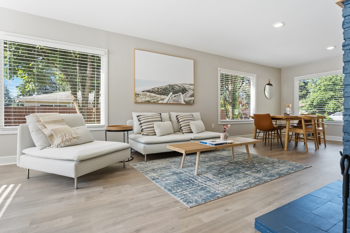 A bright living and dining area features large windows that invite natural light. A comfortable sectional sofa sits alongside a wooden coffee table and area rug, while a dining table accommodates several chairs. Neutral tones and simple decor create a serene environment.