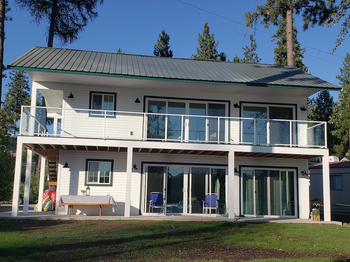 A two-story cottage features a white exterior with a green metal roof. Large glass doors on both levels allow for ample natural light. A spacious deck with railing is visible on the upper level, while the lower level includes a patio area with seating.