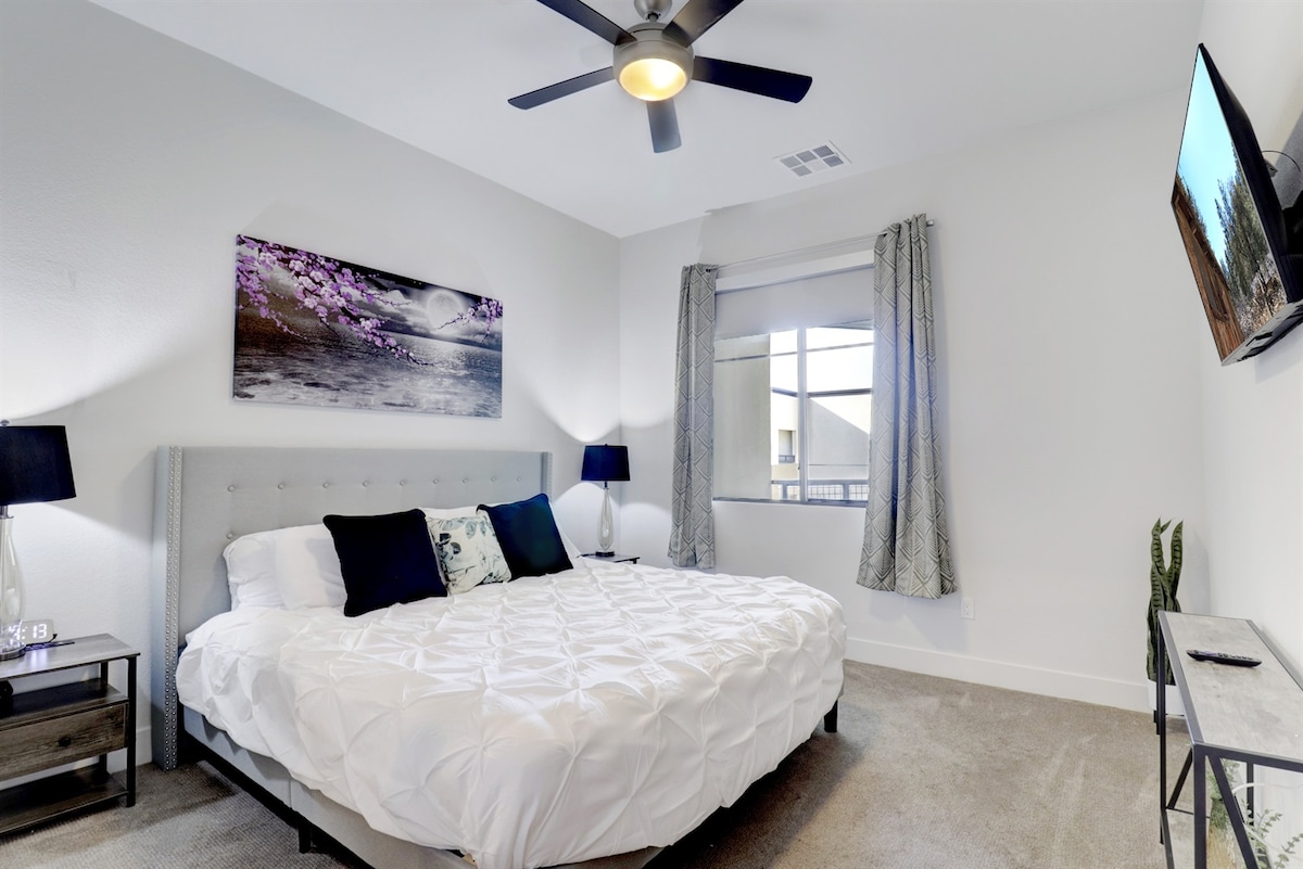 A light-filled bedroom features a spacious bed with a white quilted cover and accent pillows. Wall art depicting a serene landscape hangs above the bed. Two bedside lamps provide soft lighting, while a sleek dresser and a wall-mounted TV are positioned on opposite sides of the room.