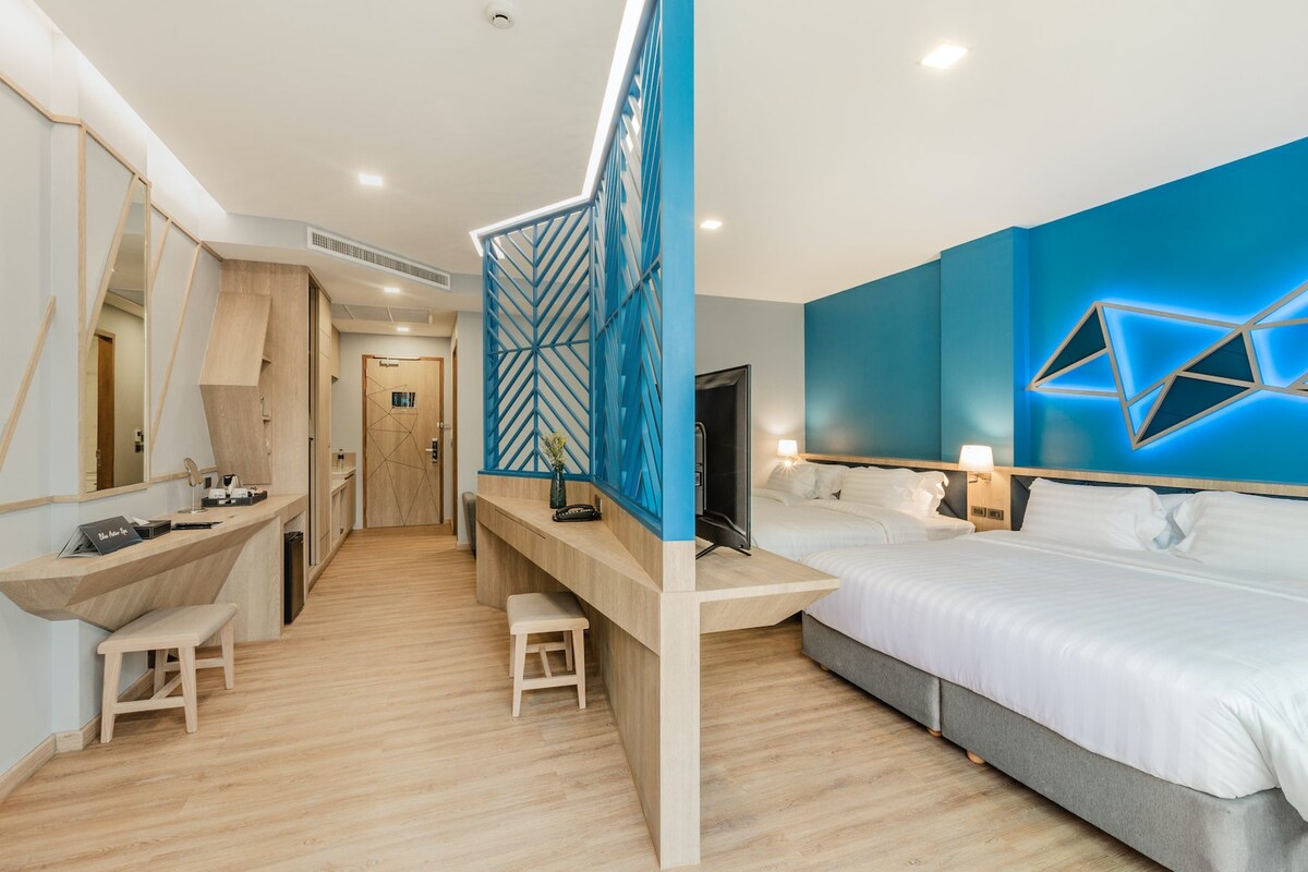 The room features a modern layout with distinct areas for relaxation and functionality. A spacious bed sits adjacent to a two-twin bed setup, and a 50-inch LED TV is visible. Light wood flooring and vibrant blue walls create a fresh ambiance.