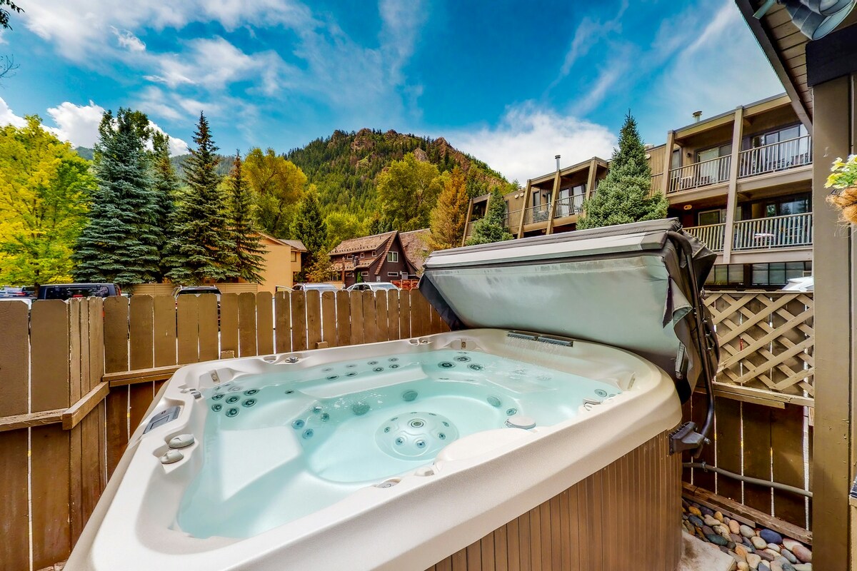 An outdoor hot tub is situated on a private patio, surrounded by a wooden fence. Lush greenery and trees are visible in the background, with mountains rising in the distance beneath a partly cloudy sky.