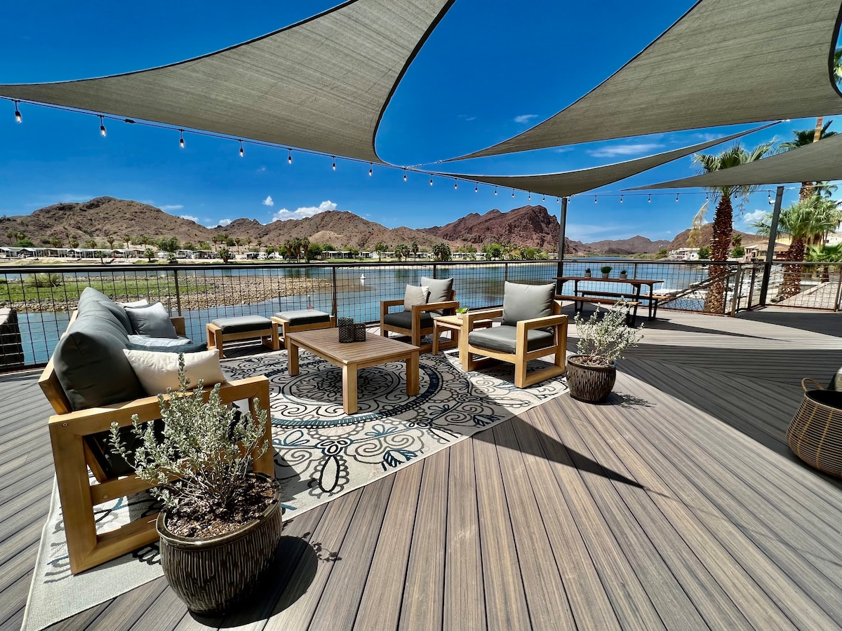 A spacious outdoor deck features modern wooden seating arrangements and a large area rug, with decorative potted plants. Shade sails create a comfortable ambiance above, while scenic river views and distant mountains provide a serene backdrop.