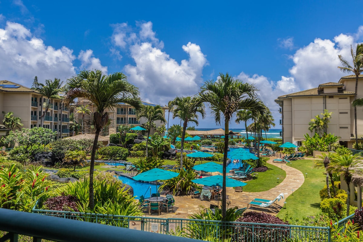 A vibrant resort landscape reveals lush tropical gardens, featuring a winding pathway that leads through colorful umbrellas and sun loungers by the pool. Surrounding buildings offer a backdrop of modern architecture, while palm trees sway gently in the breeze under a bright blue sky.