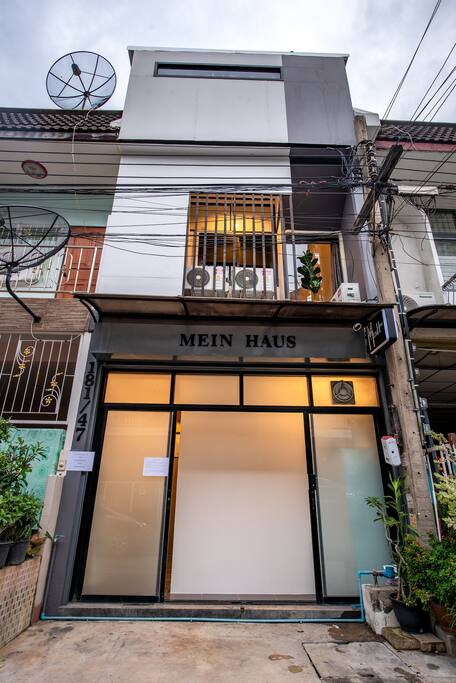 A modern townhouse exterior is presented with a minimalistic design. Large glass doors lead into the entryway, framed by a clean white façade. An upper balcony is visible, adorned with potted plants, while satellite dishes and electrical wires add urban character.