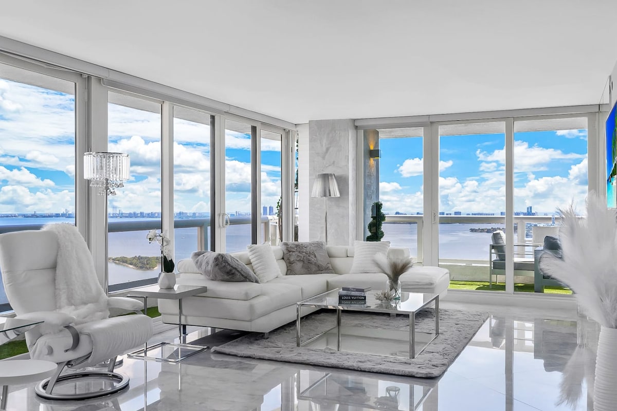 A modern living area features a spacious white sectional sofa adorned with neutral-toned cushions. Floor-to-ceiling windows provide panoramic views of the bay, allowing natural light to fill the room. A sleek coffee table sits on a soft rug, complementing the minimalist decor.