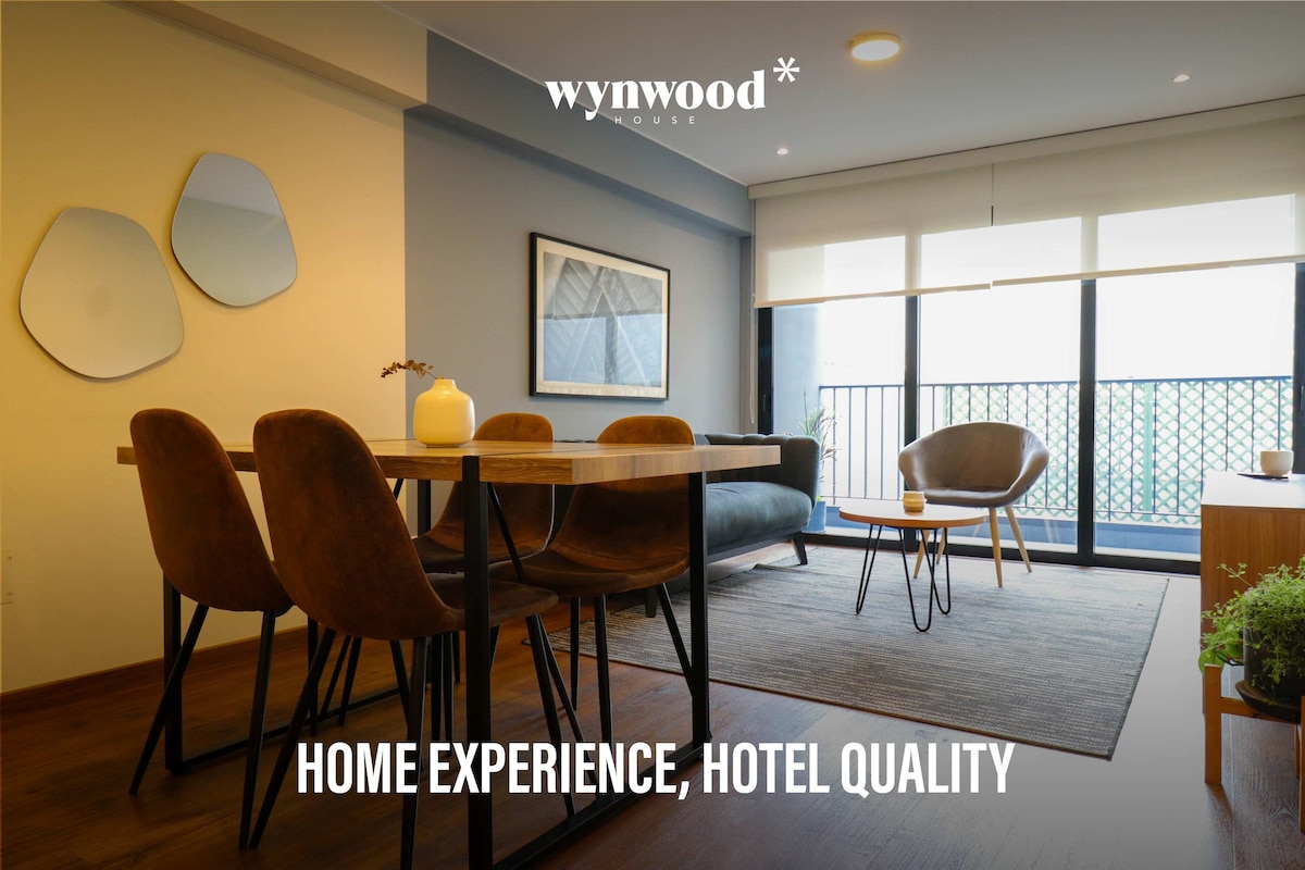 A modern living space is depicted, featuring a dining table with four brown chairs. A comfortable chair is positioned near a large window, allowing natural light to fill the room. Minimalist artwork adorns the walls, enhancing the overall decor.