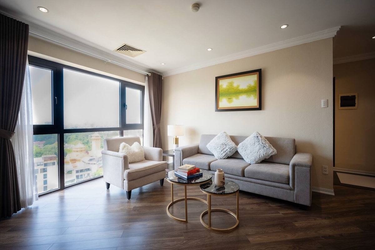 A modern living area features a spacious sofa and a stylish armchair, arranged around a simple coffee table. Large windows allow natural light to fill the room, offering views of the surrounding greenery. Neutral tones are complemented by soft decorative pillows.