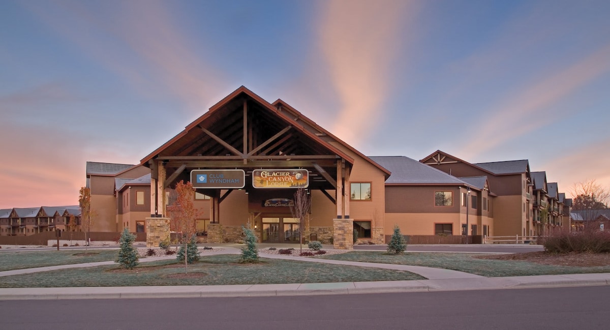 The exterior of the resort is highlighted by a spacious entrance featuring wooden beams and stone accents. Lush green landscaping and small trees frame the entrance, while soft colors in the sky suggest an early morning or late afternoon. The building’s design embraces a rustic yet modern aesthetic.