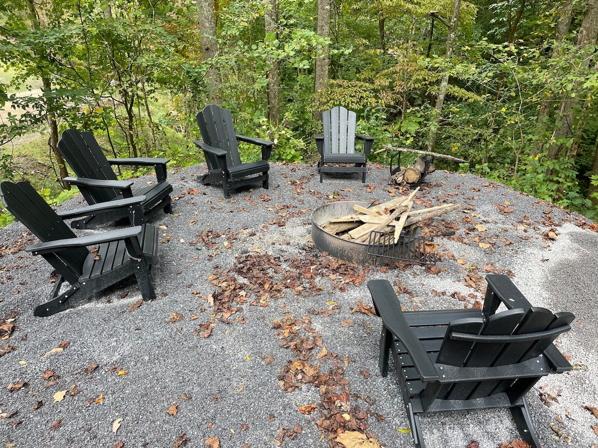 A gravel clearing surrounded by trees features multiple black Adirondack chairs arranged around a fire pit. The area showcases natural foliage with fallen leaves, creating a cozy outdoor gathering space designed for relaxation and conversation.